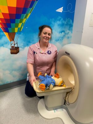 A chemo duck stuffed toy in a mini model of a hospital scanner, demonstrated by a member of hospital staff